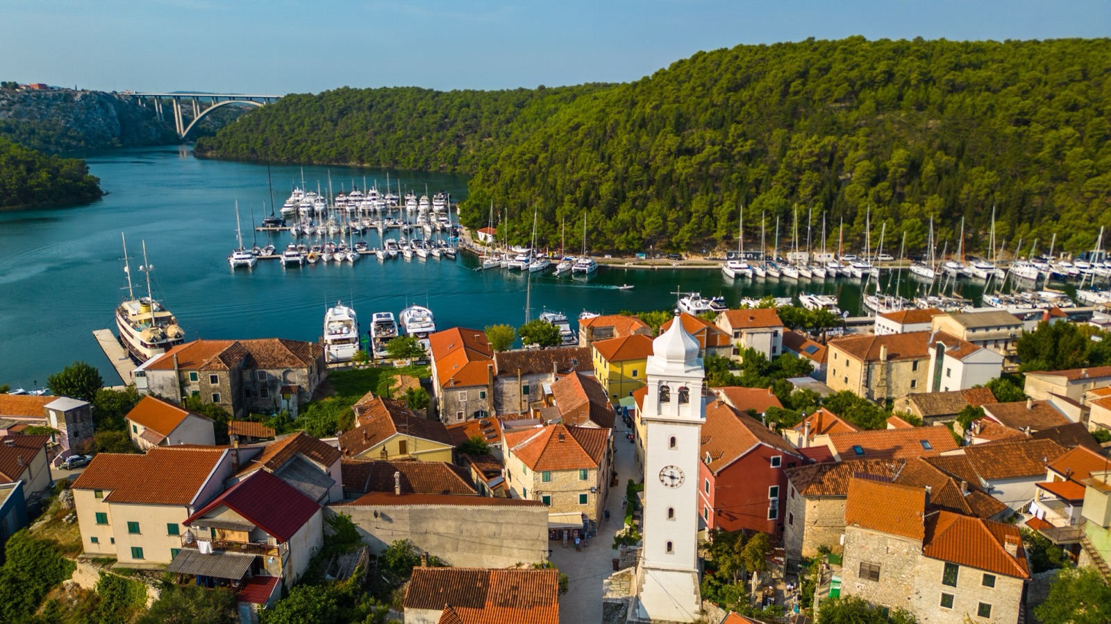 Aerial view of Skradin's red roofs and bell tower above the marina, with an arched bridge over the river beyond